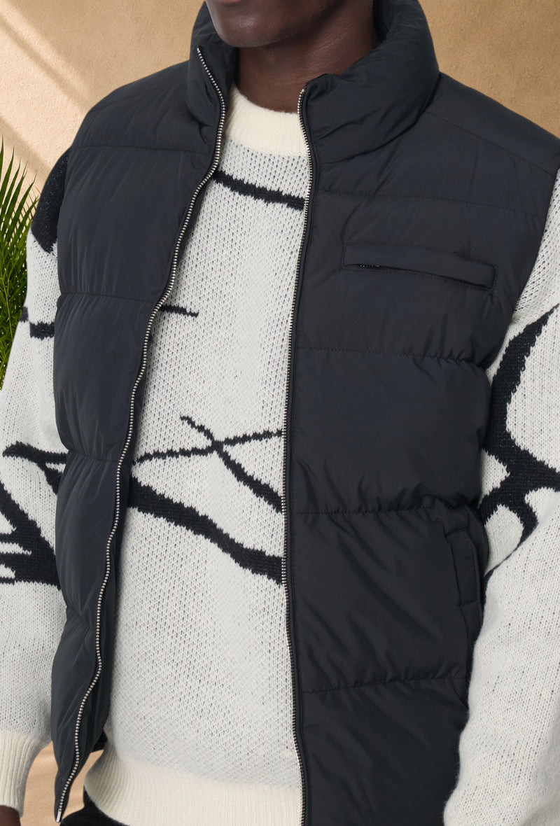 Stayin black sleeveless puffer vest with a high collar and front zipper worn over a patterned knit sweater