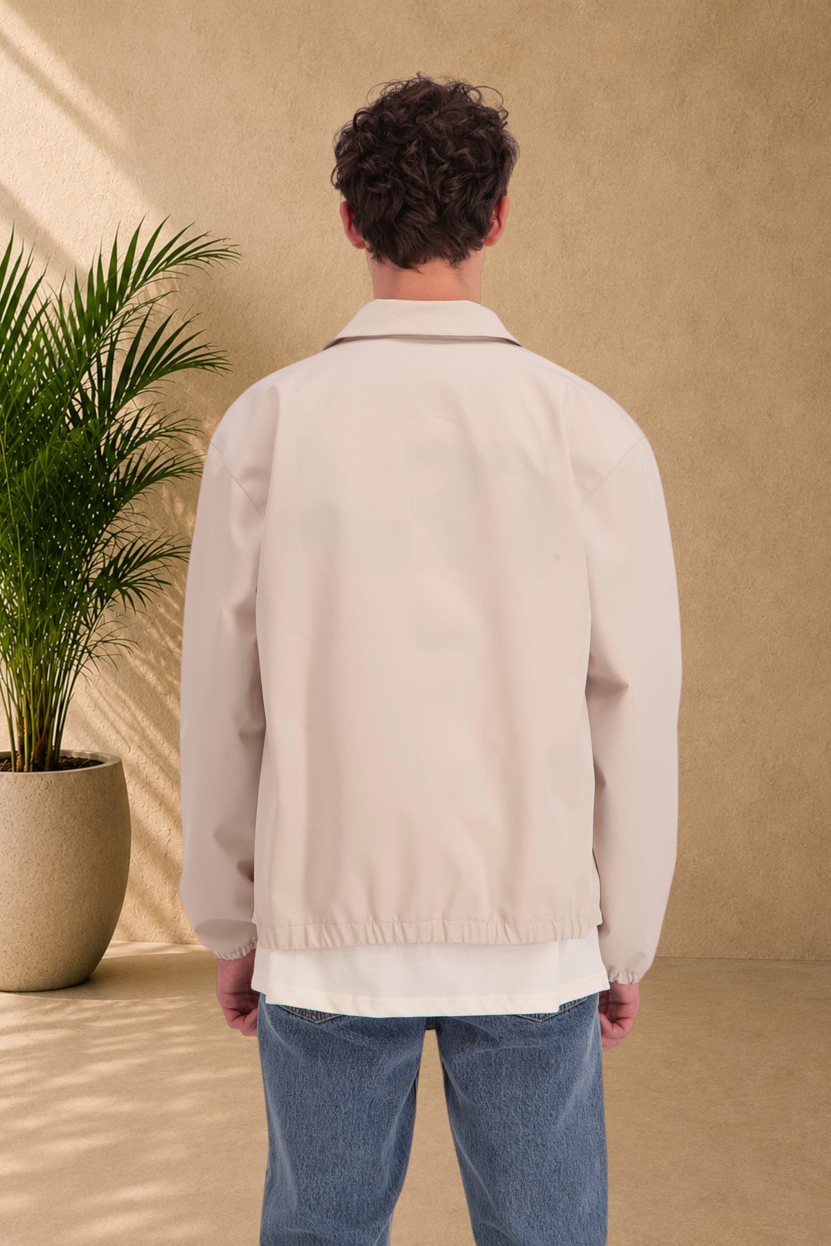 Back view of a man wearing a beige Stayin zip-up jacket with a collar and fine material, paired with blue jeans.