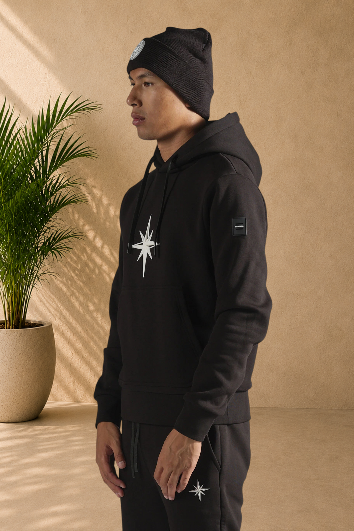 Stayin black doublehood sweatshirt with white star logo, worn by a model in a studio setting with a potted plant.